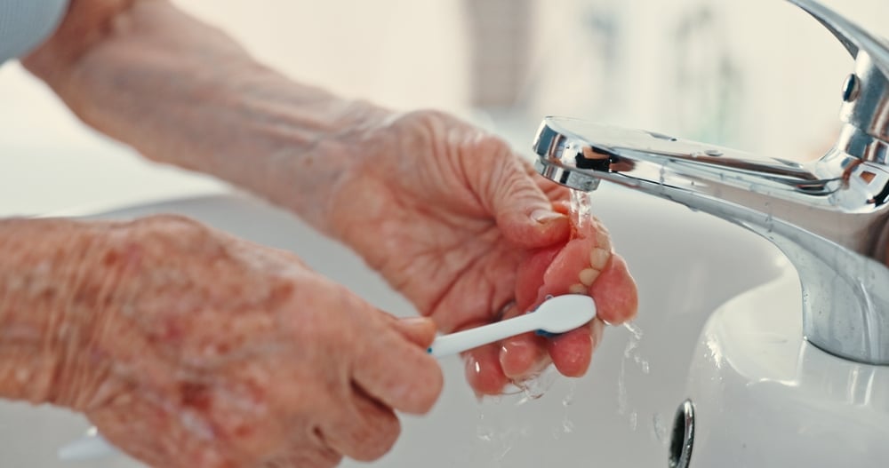 Older adult cleaning a removable denture with a toothbrush at the sink